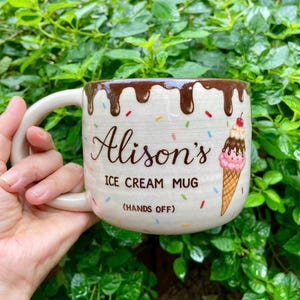 May include: A cream-coloured ceramic mug featuring a brown chocolate drip design at the rim and colourful sprinkles. The text reads Alison's ICE CREAM MUG (HANDS OFF) with a graphic of a strawberry and chocolate ice cream cone on the side.