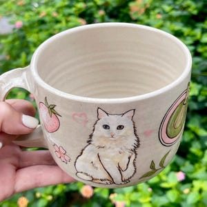 May include: Cream-colored ceramic mug with a white cat illustration. The mug features a handle and is decorated with a strawberry, flowers, and a green avocado design. Pink hearts accent the design.