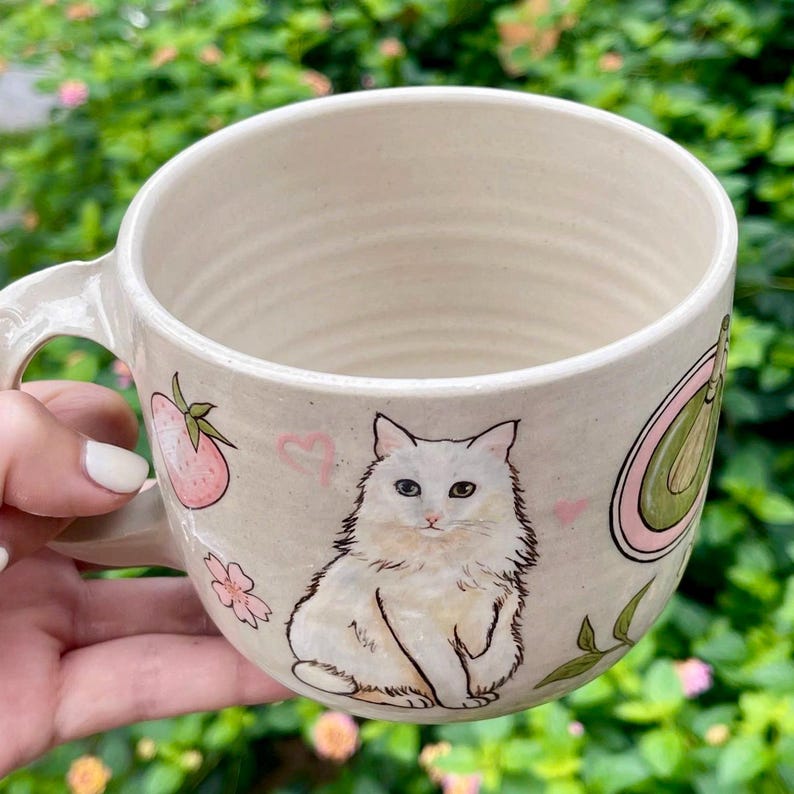 May include: Cream-colored ceramic mug with a white cat illustration. The mug features a handle and is decorated with a strawberry, flowers, and a green avocado design. Pink hearts accent the design.