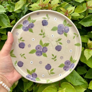 May include: A round ceramic plate with a speckled beige base featuring a hand-painted pattern of purple blueberries, green leaves, and small white flowers scattered across the surface. The plate has a smooth, glossy finish and a raised rim.
