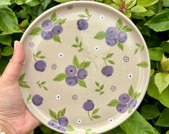 May include: A round ceramic plate with a speckled beige base featuring a hand-painted pattern of purple blueberries, green leaves, and small white flowers scattered across the surface. The plate has a smooth, glossy finish and a raised rim.