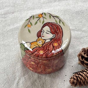 May include: A round ceramic trinket box with a lid featuring a hand-painted illustration. The lid depicts a woman with long red hair cradling a sleeping orange tabby cat. The box's base is decorated with a floral pattern.