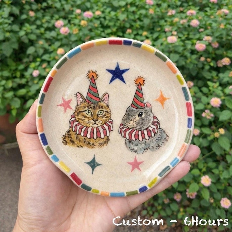 K&ouml;nnte beinhalten: Kleine, runde Keramikschale mit cremefarbenem Boden. Die Schale hat einen bunten Rand und Illustrationen einer Katze und eines Kaninchens mit Partyh&uuml;ten und R&uuml;schenkragen. Der Text "Custom - 6Hours" steht unten.