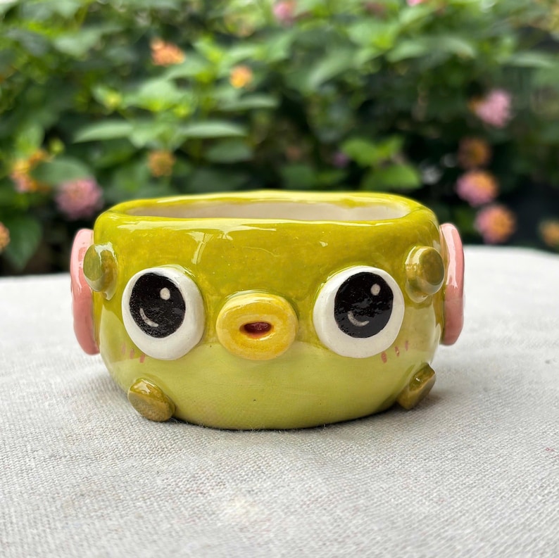 May include: A small, lime green ceramic planter shaped like a creature with large, round white eyes featuring black pupils, a small yellow circular mouth, and pink side accents. The surface is glossy, and the base has small, rounded protrusions.