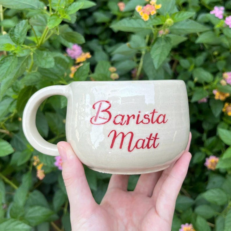Puede incluir: Taza de cer&aacute;mica color crema con forma redondeada y un asa grande. La taza presenta el texto rojo "Barista Matt" en la parte delantera. La taza se sostiene frente a un fondo de hojas verdes y flores coloridas.