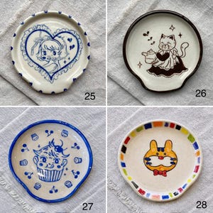 K&ouml;nnte beinhalten: Vier Keramik-L&ouml;ffelablagen mit einzigartigen Designs. Eines zeigt ein blaues Herz mit einem M&auml;dchen, ein anderes eine Katze mit einer Teekanne, ein drittes einen Cupcake und das letzte einen Hasen mit einer Fliege. Jedes hat einen gebogenen Griff.