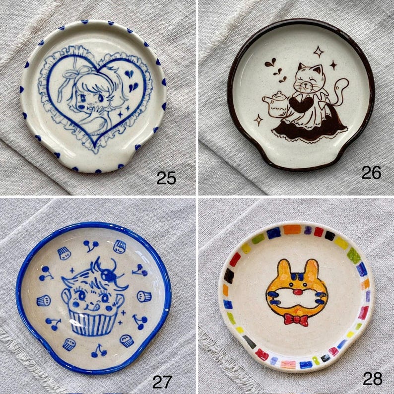 K&ouml;nnte beinhalten: Vier Keramik-L&ouml;ffelablagen mit einzigartigen Designs. Eines zeigt ein blaues Herz mit einem M&auml;dchen, ein anderes eine Katze mit einer Teekanne, ein drittes einen Cupcake und das letzte einen Hasen mit einer Fliege. Jedes hat einen gebogenen Griff.