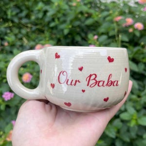 Puede incluir: Taza de cer&aacute;mica color crema con forma redondeada y un asa grande. La taza est&aacute; decorada con peque&ntilde;os corazones rojos y el texto "Our Baba" en escritura roja. La taza se sostiene en una mano sobre un fondo verde borroso.