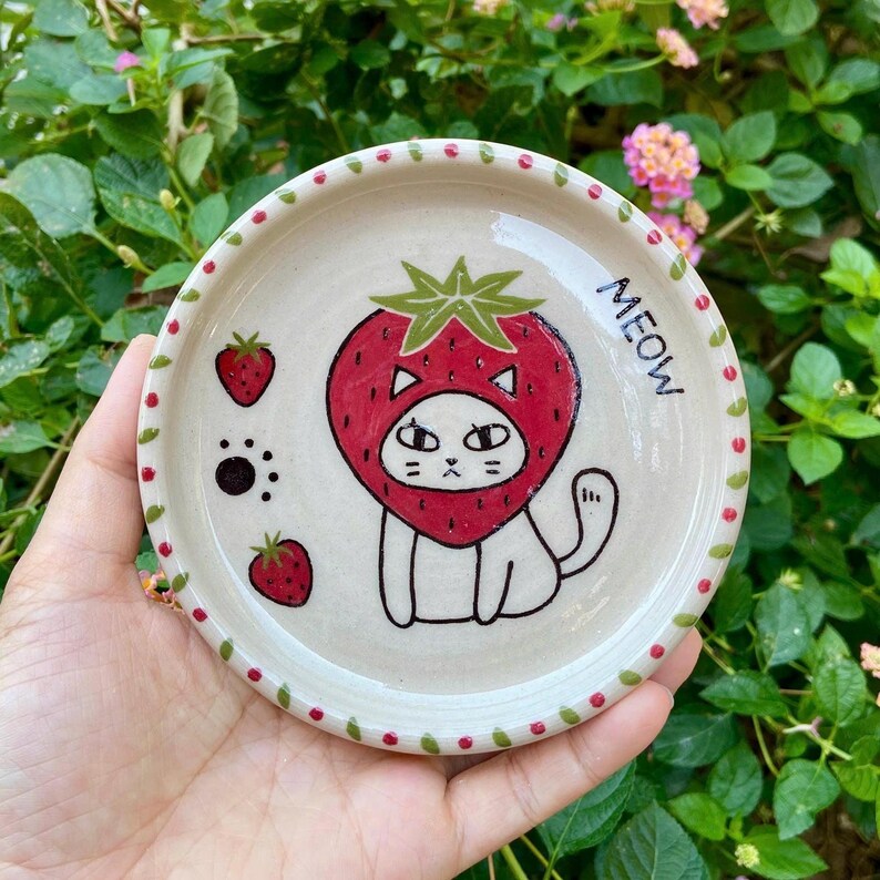 May include: A round ceramic dish with a cream-colored base and a red dotted rim. The center features a hand-drawn illustration of a white cat wearing a strawberry costume, two small strawberries, a paw print, and the word MEOW in black ink.