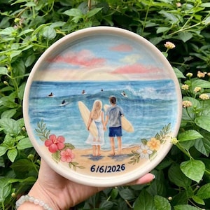 May include: A round ceramic plate featuring a hand-painted beach scene with two people holding surfboards, facing the ocean. The design includes pink clouds, blue waves, and colorful hibiscus flowers at the bottom, with the date 6/6/2026 printed below.