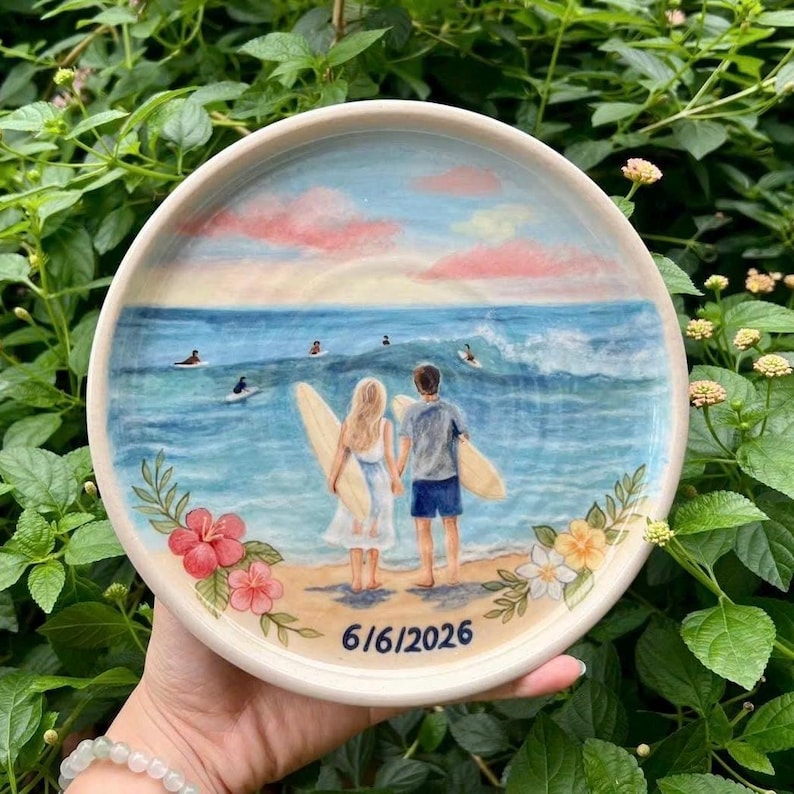 May include: A round ceramic plate featuring a hand-painted beach scene with two people holding surfboards, facing the ocean. The design includes pink clouds, blue waves, and colorful hibiscus flowers at the bottom, with the date 6/6/2026 printed below.
