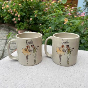 May include: Two beige ceramic mugs with handles. Each mug features the word "Copp&eacute;lia" in black script and a ballet-themed illustration of two dancers in yellow and green attire. The mugs are set on a white surface.