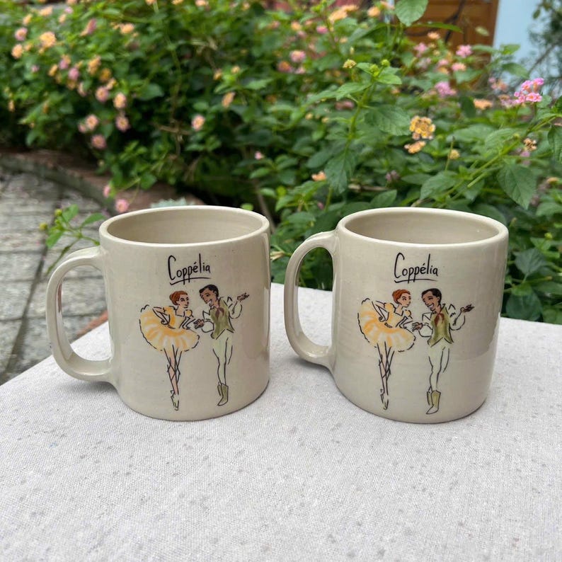 May include: Two beige ceramic mugs with handles. Each mug features the word "Copp&eacute;lia" in black script and a ballet-themed illustration of two dancers in yellow and green attire. The mugs are set on a white surface.