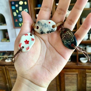 May include: Three ceramic bracelets are displayed. One has red floral designs on a white oval. Another features a mountain landscape. The third has a sun design on a dark blue oval. Each bracelet is connected with a brown cord, creating a unique accessory.