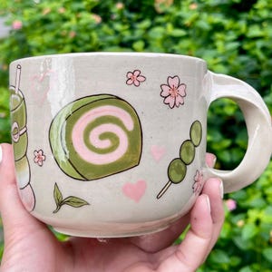 May include: A ceramic mug with a light beige finish, decorated with illustrations of Japanese sweets and drinks. The design includes a matcha drink, a green and pink swirl cake, dango, pink hearts, and cherry blossoms.