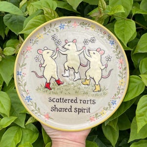 May include: A round ceramic plate with a cream-coloured surface featuring three illustrated white mice wearing colourful boots and holding white flowers. The text "scattered rarts shared spirit" is printed in black below the mice, surrounded by a floral border.