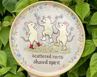May include: A round ceramic plate with a cream-colored surface featuring three illustrated white mice wearing colorful boots and holding white flowers. The text "scattered rarts shared spirit" is printed in black below the mice, surrounded by a floral border.