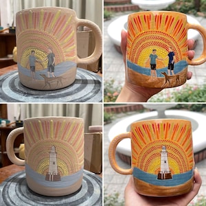 May include: Hand-painted ceramic mug featuring a sunset design with two figures and a dog on a beach. The mug has a large handle and is painted with orange, yellow, and blue hues. The design is repeated on the other side of the mug, but with a lighthouse.