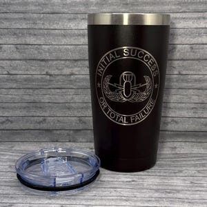 May include: Black insulated tumbler with a silver rim and a clear lid. The tumbler features the text "INITIAL SUCCESS OR TOTAL FAILURE" in a circular design with a bomb graphic.