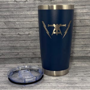 May include: A navy blue insulated tumbler with a silver rim and a clear lid. The tumbler features a silver graphic design of a stylized symbol with lightning bolts on either side. The lid is off to the side.