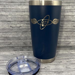 May include: A navy blue insulated tumbler with a silver rim and a clear lid. The tumbler features a gold-colored graphic design of a winged oval with stars and dots. The lid is off to the side.