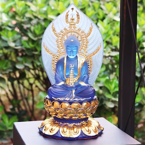 Puede incluir: Estatua de Buda de vidrio azul con halo dorado y base de loto. La estatua está sentada en posición meditativa con las manos en mudra.