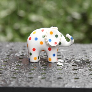 Pode incluir: Uma pequena figura de elefante de cerâmica branca adornada com bolinhas coloridas em vermelho, azul, verde e laranja. O elefante tem um colar de pérolas e um adorno de cabeça. A figura está sobre uma superfície escura e texturizada.