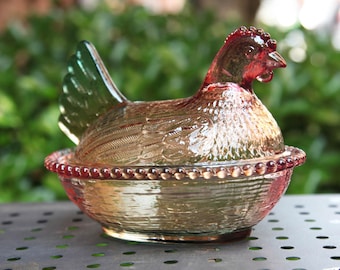 Op de afbeelding: Een decoratieve glazen schaal in de vorm van een kip met een deksel. De kip is roodbruin van kleur met een groene staart en een rode kam. De schaal heeft een textuur en staat op een metalen oppervlak.