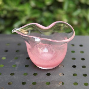 Op de afbeelding: Een kleine, roze glazen roomkannetje met een unieke, golvende rand en een tuit. Het roomkannetje heeft een afgeronde basis en een delicate, transparante uitstraling. De roze kleur is consistent, waardoor het een zachte, elegante uitstraling heeft.