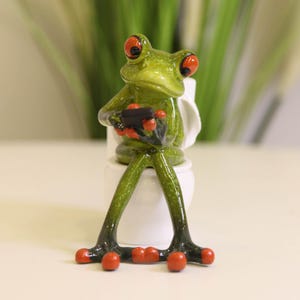 Puede incluir: Una caprichosa figura de rana verde con ojos y pies naranjas, sentada en un inodoro blanco. La rana sostiene un pequeño objeto negro. La figura tiene un acabado brillante. Artículo decorativo.