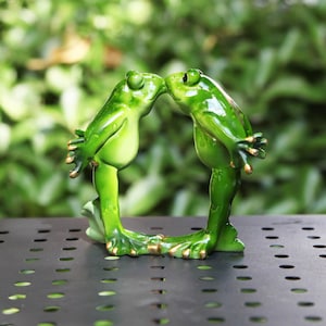 May include: A green frog figurine with two frogs kissing. The frogs are glossy and have gold accents on their hands and feet. The figurine is set against a blurred green background, suggesting an outdoor setting.