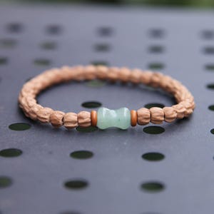 May include: A beaded bracelet with a light brown, textured band. The bracelet features a light green, hourglass-shaped bead in the center, flanked by smaller brown beads and orange spacers. The bracelet is set against a dark gray surface.