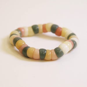 May include: A bracelet made of rounded beads in a variety of colours, including green, pink, yellow, and white. The beads are strung together to form a circular shape. The beads have a smooth, polished appearance.