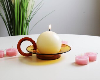 Könnte beinhalten: Eine weiße runde Kerze mit einer Flamme, die auf einem braunen Glaskerzenhalter mit rotem Griff brennt. Auf einer weißen Oberfläche befinden sich mehrere rosa Teelichter.