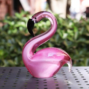 Peut inclure: Une sculpture de flamant rose en verre avec un bec noir. L'oiseau décoratif a un cou incurvé et est exposé sur une surface métallique. Le flamant rose est d'une couleur rose vif et est un objet décoratif.