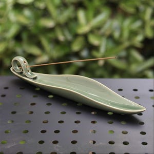 May include: A sage green ceramic incense holder with a curved design. The holder has a long, shallow dish shape with a curled end that has three small holes. A lit incense stick is placed in the holder. The background is blurred green foliage.