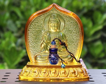 May include: A golden glass sculpture of a Buddha figure with a small blue glass bottle pendant hanging from the neck. The sculpture is on a base with intricate details.