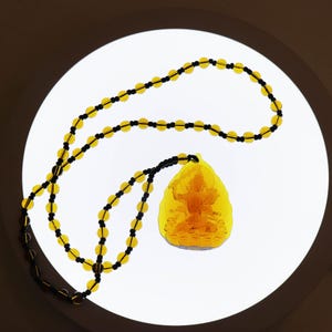 May include: A yellow amber necklace with a pendant featuring a detailed carving of a Buddha figure. The necklace is made of small, round beads.