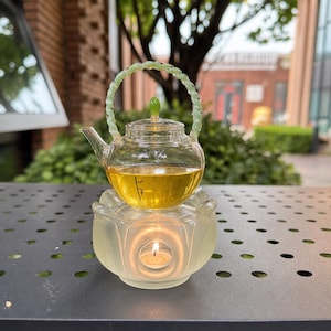 Könnte beinhalten: Eine Teekanne aus klarem Glas mit einem blassgrünen, gedrehten Griff und einem kleinen grünen Knauf auf dem Deckel, gefüllt mit goldener Flüssigkeit. Sie steht auf einem milchigen Glassockel in Lotusblütenform mit einem brennenden Teelicht darin.