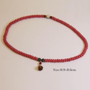May include: A pink beaded necklace with a small pendant. The necklace is made of small, round beads. The pendant is a small, round, brown charm with a dotted pattern. The necklace measures 0.9 x 0.6 cm.