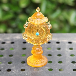 Pode incluir: Um objeto decorativo detalhado, de cor âmbar, com uma pedra central turquesa. O objeto tem um design ornamentado com uma base de pedestal e um topo em forma de cúpula. O fundo é verde desfocado, sugerindo um ambiente ao ar livre.