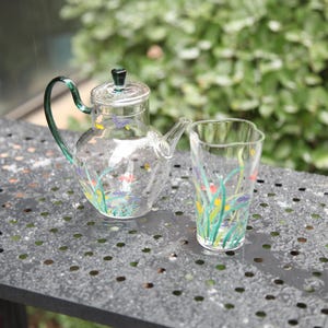 Puede incluir: Una tetera de cristal transparente y un vaso a juego con un diseño floral. La tetera tiene un asa y tapa verdes, y el vaso tiene una parte superior ondulada. El diseño floral incluye mariposas, flores y hierba.