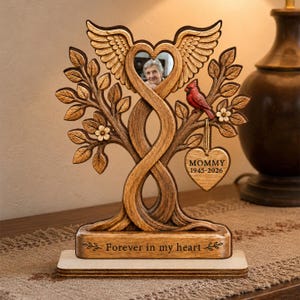 May include: A wooden memorial plaque featuring a tree with intertwined trunks forming a heart shape at the top, adorned with angel wings, a red cardinal, and a hanging heart tag inscribed with MOMMY 1945-2026. The base reads Forever in my heart.
