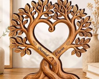 Op de afbeelding: Een houten sculptuur met twee verstrengelde bomen die in het midden een hartvormige lege ruimte vormen. De stammen zijn gegraveerd met de namen Daniel en Olivia, en er is een klein hartsymbool in het hout bij de namen uitgesneden.