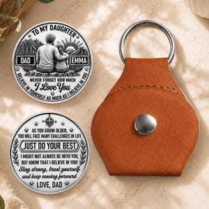 May include: Two circular silver-toned metal coins with black engraved text and imagery, accompanied by a tan leather keychain pouch with a silver metal ring. One coin depicts a parent and child with the text TO MY DAUGHTER DAD EMMA NEVER FORGET HOW MUCH I LOVE YOU BELIEVE IN YOURSELF AS MUCH AS I BELIEVE IN YOU. The second coin features the text AS YOU GROW OLDER, YOU WILL FACE MANY CHALLENGES IN LIFE JUST DO YOUR BEST I MIGHT NOT ALWAYS BE WITH YOU, BUT KNOW THAT I BELIEVE IN YOU! Stay strong, trust yourself and keep moving forward LOVE, DAD.