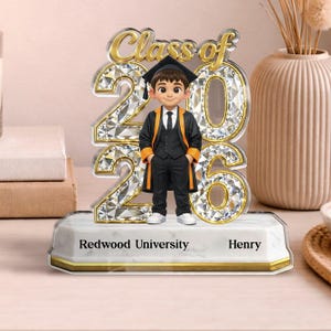 May include: An acrylic graduation ornament with a cartoon graduate in cap and gown, standing before the words "Class of 2026." The numbers are decorated with a diamond pattern. The base reads "Redwood University" and "Henry."