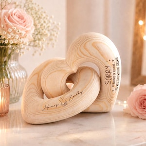 May include: A light-colored wooden sculpture featuring two interlocking hearts. The wood grain is visible throughout. Engraved text reads "Henry & Emily" with a small hand-holding icon, and "SORRY NO REFUNDS OR RETURNS... YOU'RE STUCK WITH ME FOREVER".