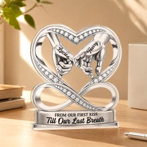 Puede incluir: Un adorno de acrílico transparente en forma de corazón con dos manos plateadas entrelazadas por los dedos meñiques. El diseño incluye cristales incrustados, los nombres Lucie y Harry, y una base con el texto FROM OUR FIRST KISS Till Our Last Breath.