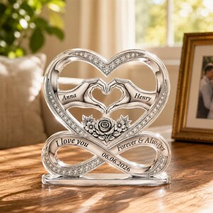 May include: A silver-toned heart-shaped ornament featuring two hands forming a heart, a central rose, and an infinity base. Engraved text includes "Anna," "Henry," "I love you," "Forever & Always," and the date "06.06.2026" with crystal accents.