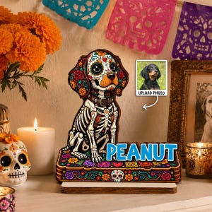 May include: A wooden dog figurine featuring a colourful sugar skull pattern, a collar, and the name PEANUT in blue letters on a decorative base. A small square inset shows a photo of a black and tan dog with the text UPLOAD PHOTO below it.